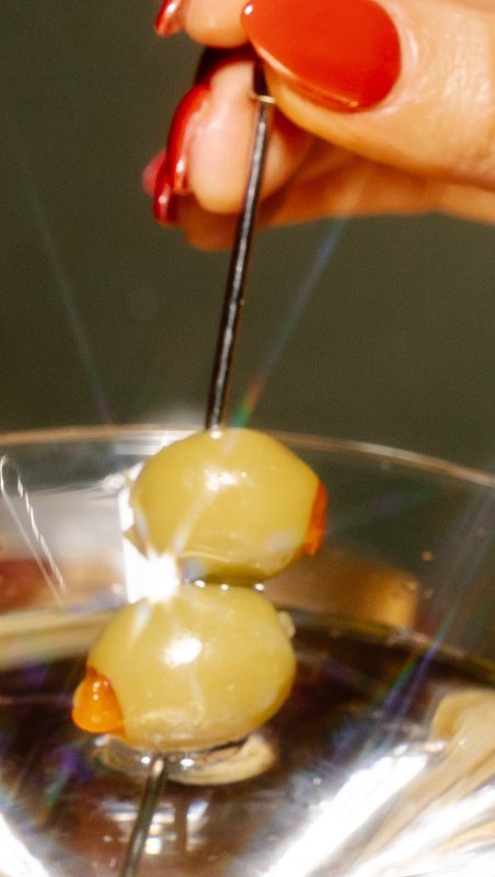 A hand with red nails lifting olives from a martini